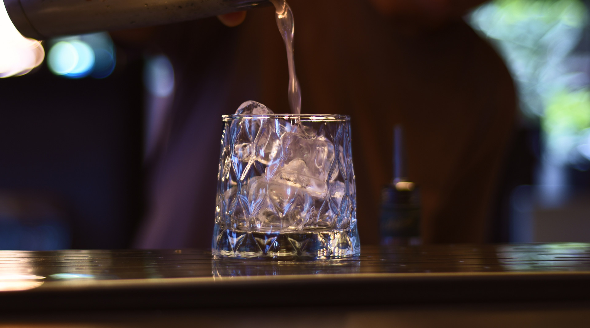 Un bicchiere basso con ghiaccio cristallino, mentre il drink viene versato.
Vetro lavorato, riflessi nitidi, banco in acciaio. Lo sfondo resta sfocato, il focus è tutto sul bicchiere.