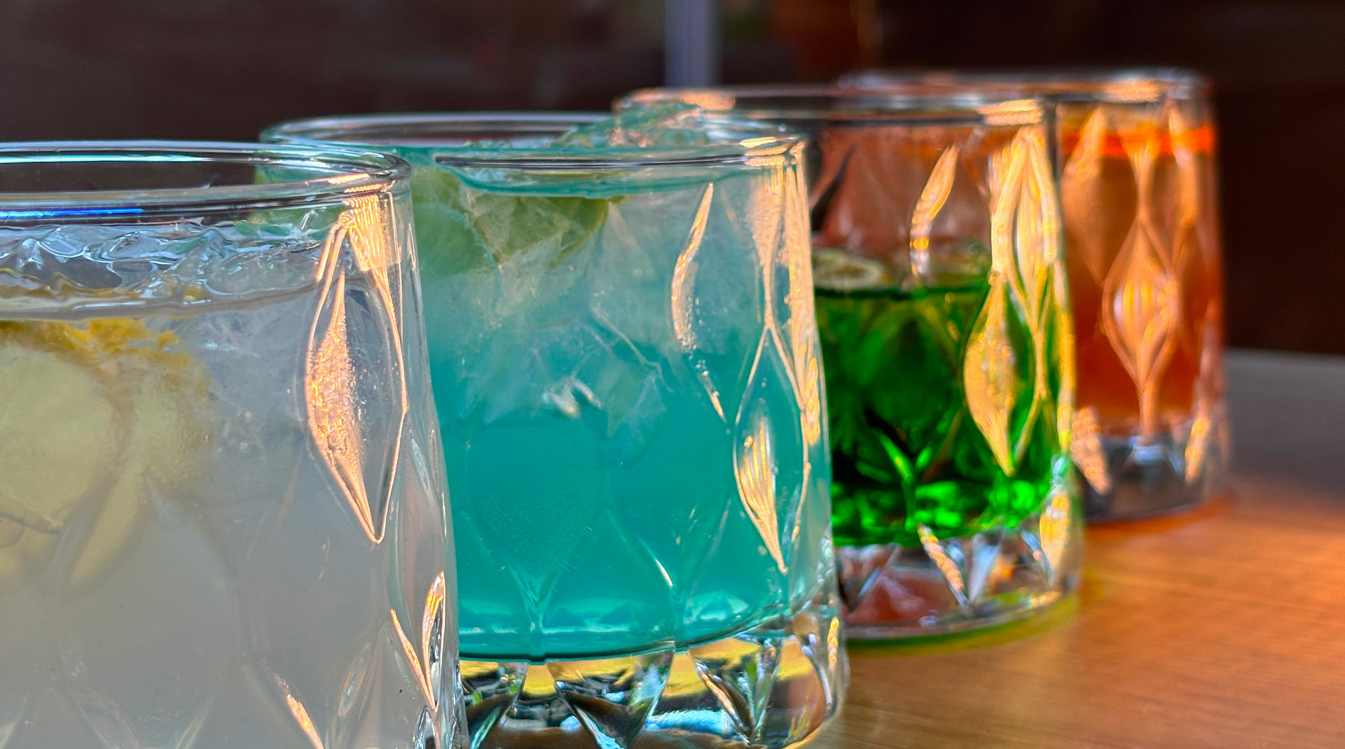 Quattro cocktail colorati serviti in tumbler lavorati, disposti su un piano in legno.
Colori accesi, ghiaccio e guarnizioni fresche, con luce naturale che valorizza i riflessi del vetro.