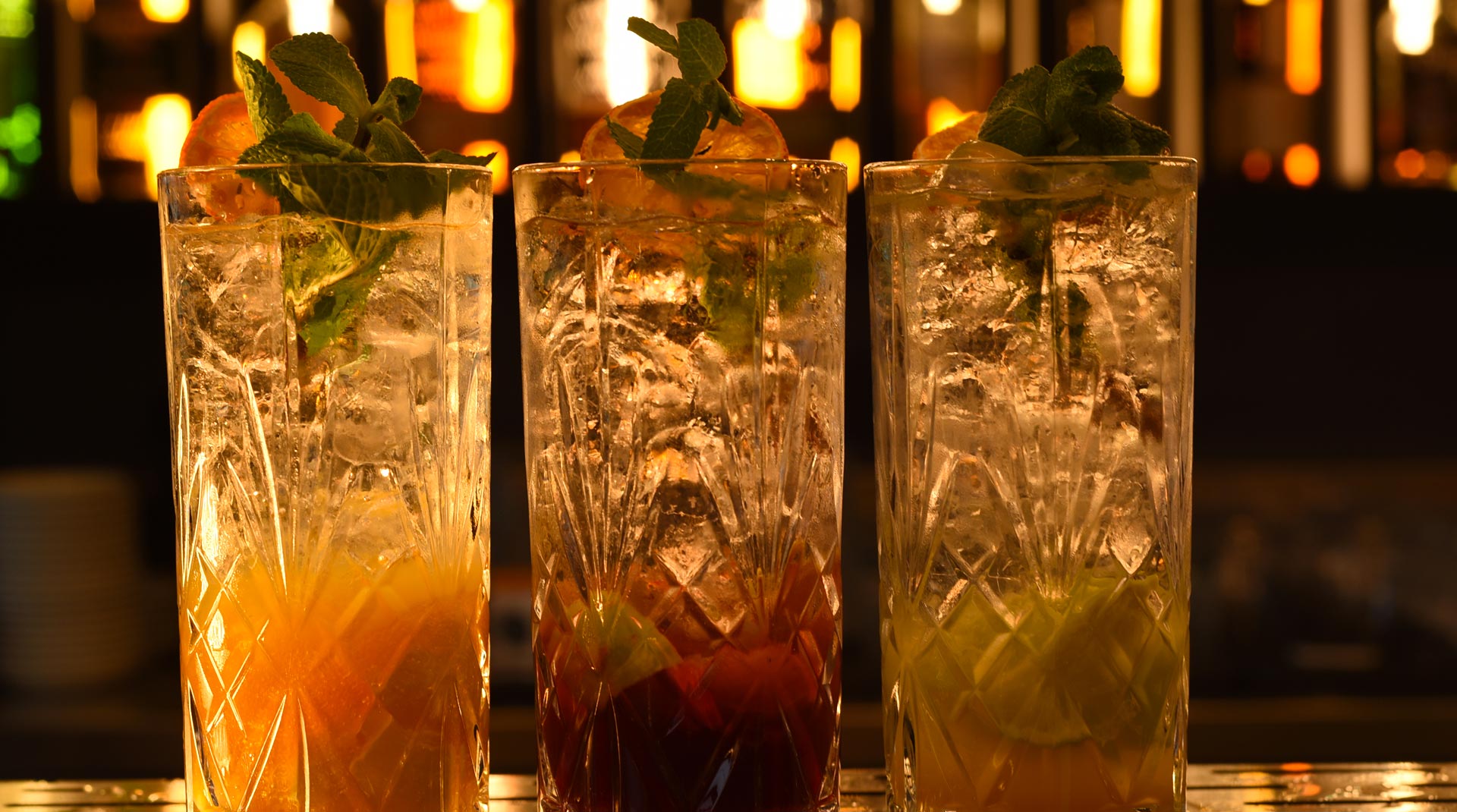 Tre highball in vetro inciso, riempiti con ghiaccio e mix stratificati. Ogni drink è guarnito con menta fresca e una fetta di agrume essiccato. Luce calda dal retrobanco crea riflessi dorati e valorizza la trasparenza dei bicchieri.