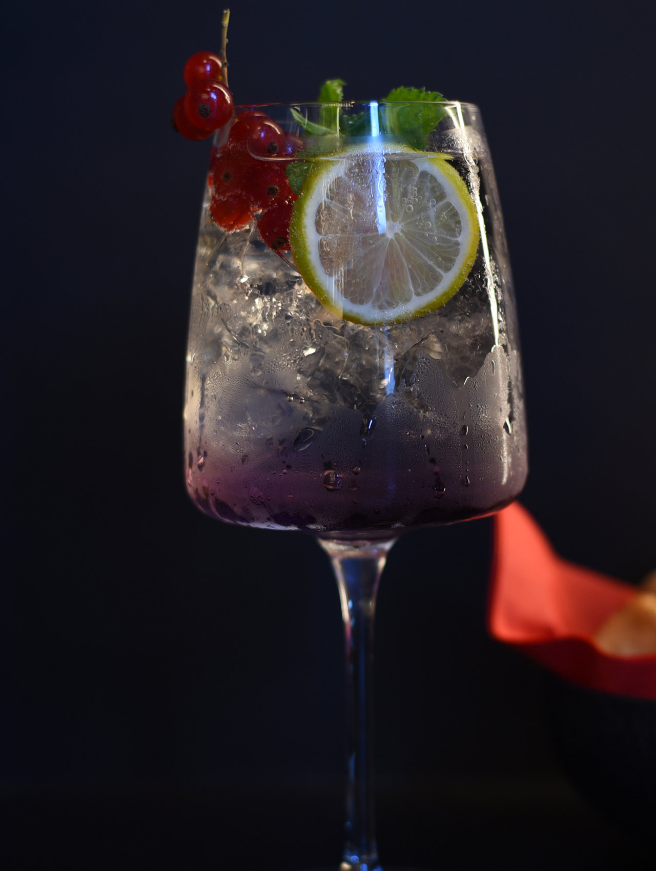 Drink servito in un calice ampio con ghiaccio abbondante. Base viola sfumata, guarnito con ribes rosso, fetta di lime e foglie di menta. Condensa visibile sul vetro, sfondo scuro e pulito con un tocco di colore laterale.