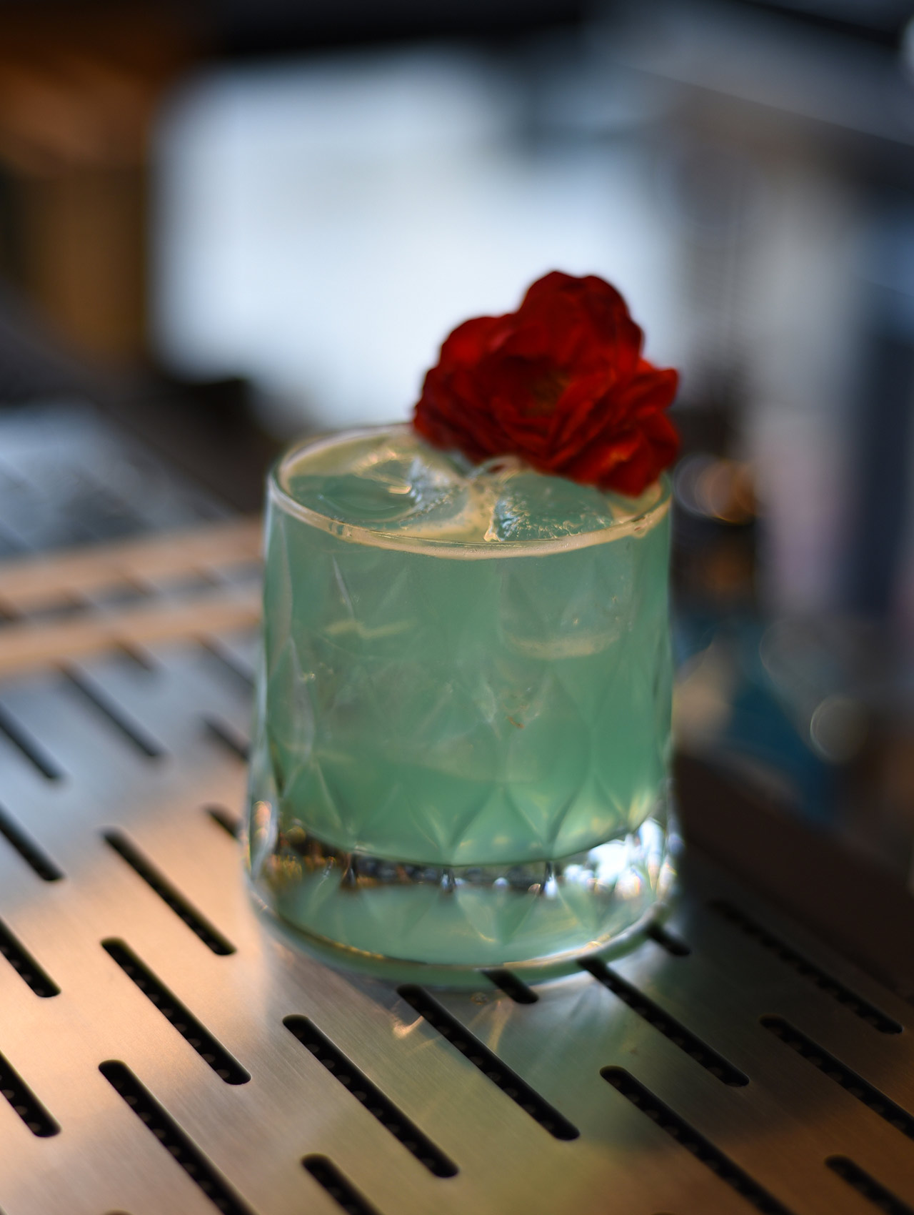 Cocktail servito in un tumbler basso con vetro lavorato. Il drink ha una colorazione verde acqua, con ghiaccio e guarnizione floreale rossa. La superficie in acciaio lucido riflette luci soffuse dell’ambiente.